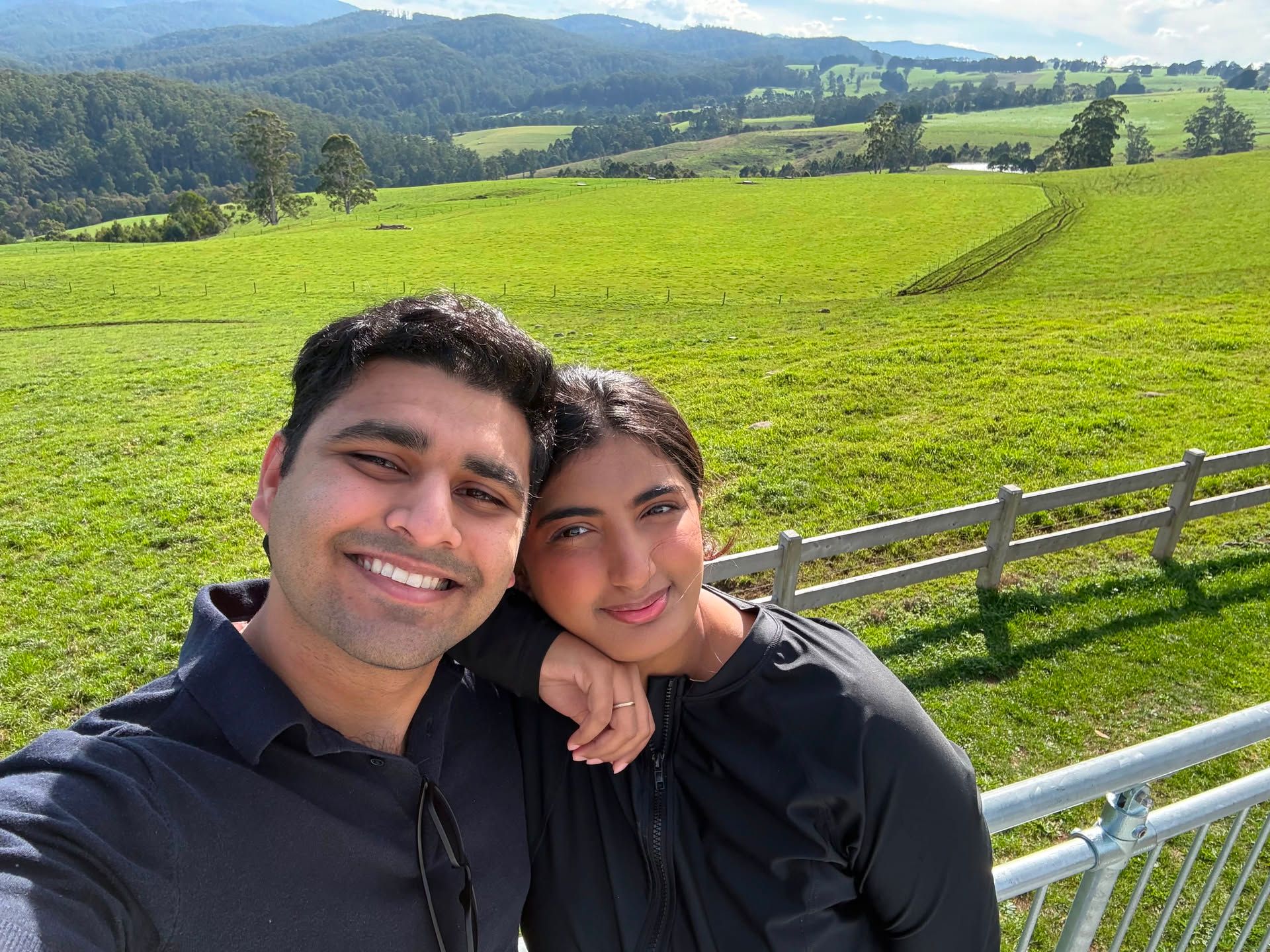 Faisal Karim and Fatima Parkar smiling together in front of green rolling hills and a farm fence